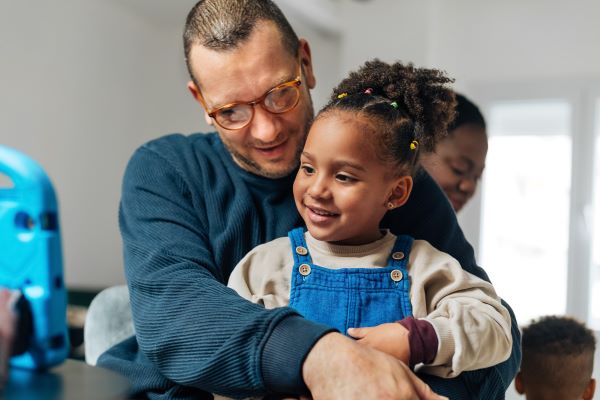 A man wearing a dark blue jumper hugs a small girl who is wearing denim dungarees and watching something on a tablet. In the background you can see another young child and a woman who is watching him.