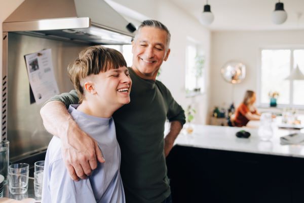 A man and teenage boy are stood in a kitchen. The smiling man, wearing a dark grey top has his arm around the teenage boy who is wearing a grey top. In the background you can see a kitchen counter and a woman sat at a table.