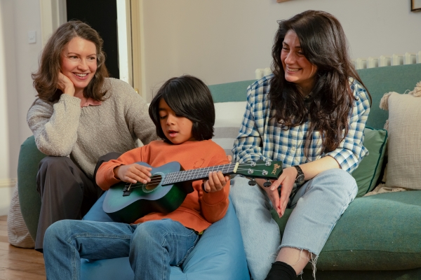 A young child in an orange jumper and blue jeans is playing a dark green guitar whilst sat on a blue beanbag. In the background are two women sat on a sofa smiling and watching on. One on the left is wearing a grey jumper and the other is wearing a checked shirt.