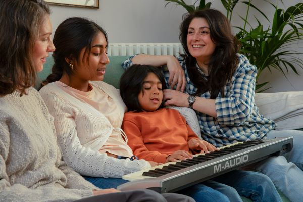 Two women and two children are sat on a sofa, with one of the children playing a keyboard. The woman on the left is wearing a cream jumper, a teenage child is sat on her right also wearing a cream top. The young child playing the keyboard is wearing an orange top, and the woman on the right of the sofa is wearing a check blue top.