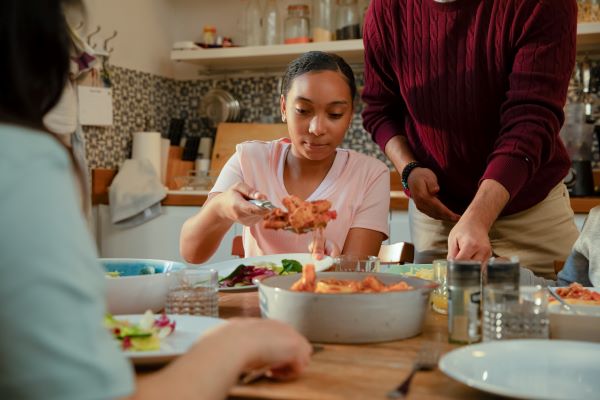 A girl in a pink t shirt is portioning some food out of a bowl onto her plate. Stood next to her is someone in a burgundy jumper and another person watches on in the foreground. In the background you can see they are in a busy home kitchen with a range of jars and utensils.