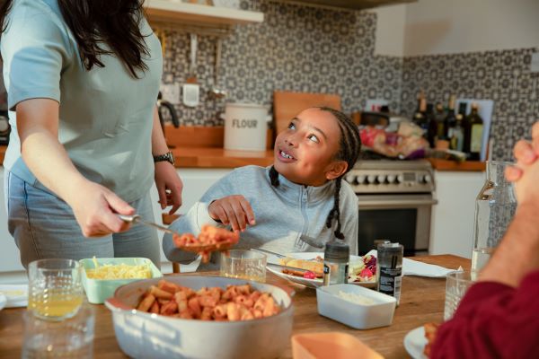 In the centre of the photo a young girl with dark hair looks up and smiles at a woman in a grey top who is passing her some pasta. In the foreground is the clasped hands of someone watching on. They are all around a kitchen table with pasta, cheese, drinks and condiments on. In the background is a kitchen work surface.