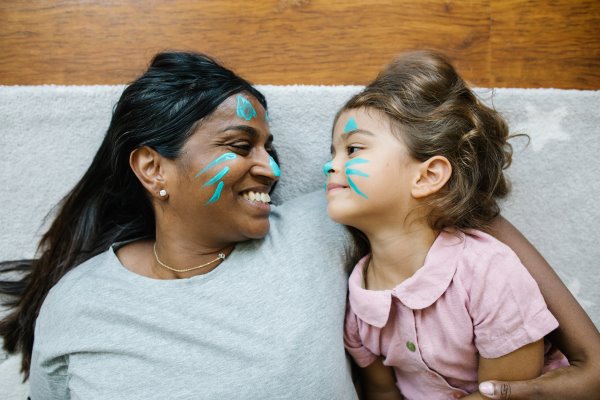 A woman in a grey t shirt is lying on a blanket smiling at a small girl with dark hair in a pink top. Both have blue face paint on.