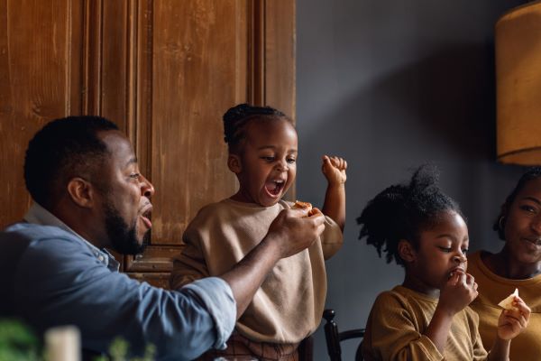 A man with a blue top holds some food out in his hand for a young girl in a beige top to eat. To there right, an older girl is sat on a chair eating a crisp, and to her right a woman watches her and smiles.