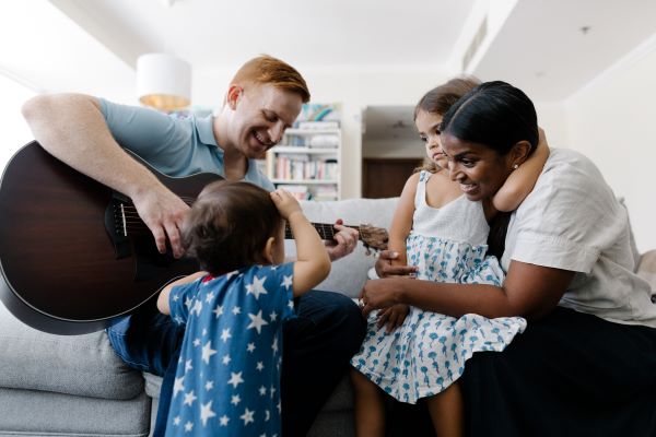 A man with ginger hair and a blue top is sat on a sofa playing a guitar. A small child is stood in front of him and is wearing a blue top with white stars. To the man's right is a woman with black hair with a white top and dark trousers who has a young girl with a white and blue dress sat on her lap.