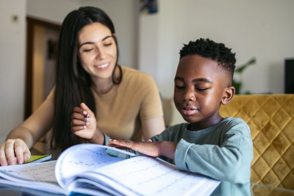 A women in a beige jumper is helping a young boy do his homework. He is wearing a blue top who is sat on a mustard chair.