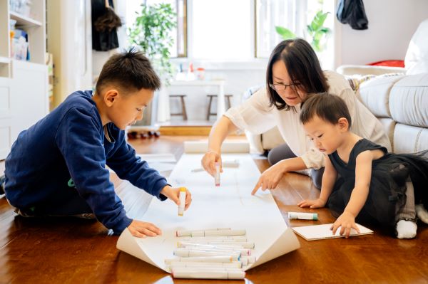 In a lounge a young boy in a dark blue top and a small girl in a dress are colouring on a long roll of paper. A woman with glasses and wearing a white top is sat behind the young child pointing at the paper holding a colouring pen. In the background you can see a cream sofa and a table with some houseplants.