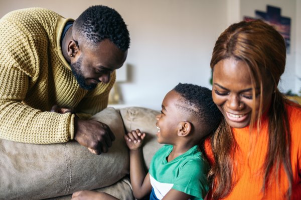 A woman in an orange top is sat on a brown sofa with a young boy next to her. A man in a beige jumper is leaning over the sofa and they're smiling at each other.