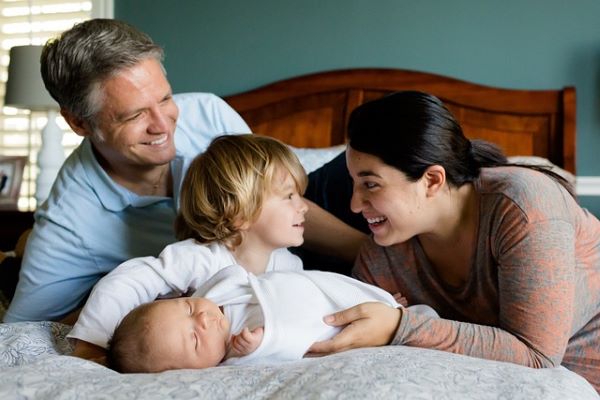 A man in a blue t-shirt smiles on whilst a woman with dark hair and a red and grey top smiles at a young boy in a white tshirt. Whilst a young baby sleeps on a grey bedding. In the background is a dark grey headboard.