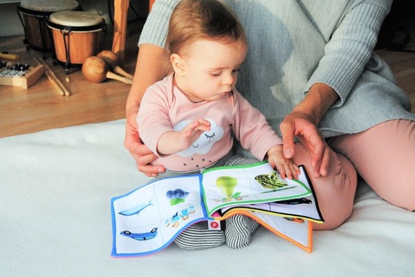 A person in a grey jumper and pink trousers is sat reading a book which has pictures on to a baby in a pink jumper with a cloud and smiley face on. In the background are some musical instruments.