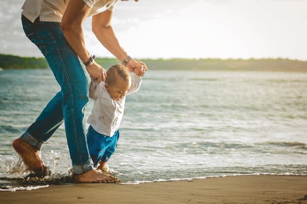 A barefoot man in jeans is holding both hands of a child in a white shirt and blue trousers playing on the edge of the sea.