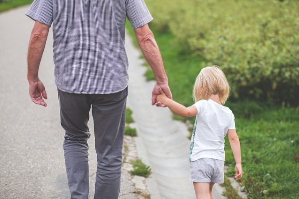 An older man wearing a grey and white checked top and dark trousers holds the hand of a young child in shorts and tshirt whilst walking along the edge of a road.