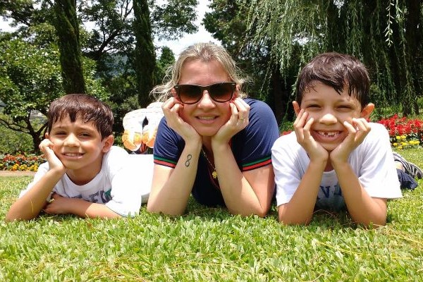 A woman in a dark blue top is lying on grass wearing sunglasses whilst two small boys, both wearing white tops smile at the camera lying either side of her.