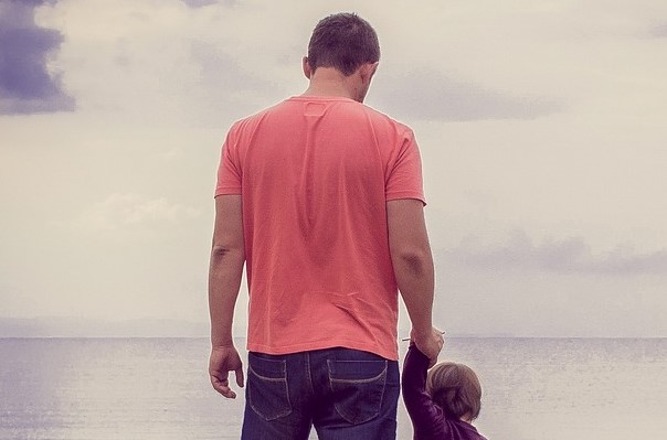 A man in a pink top holds a small child's hand whilst staring out to sea.