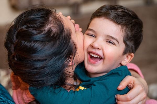 A woman with dark hair has picked up a small boy and is kissing him on the cheek. The young boy in a blue top is smiling and laughin.