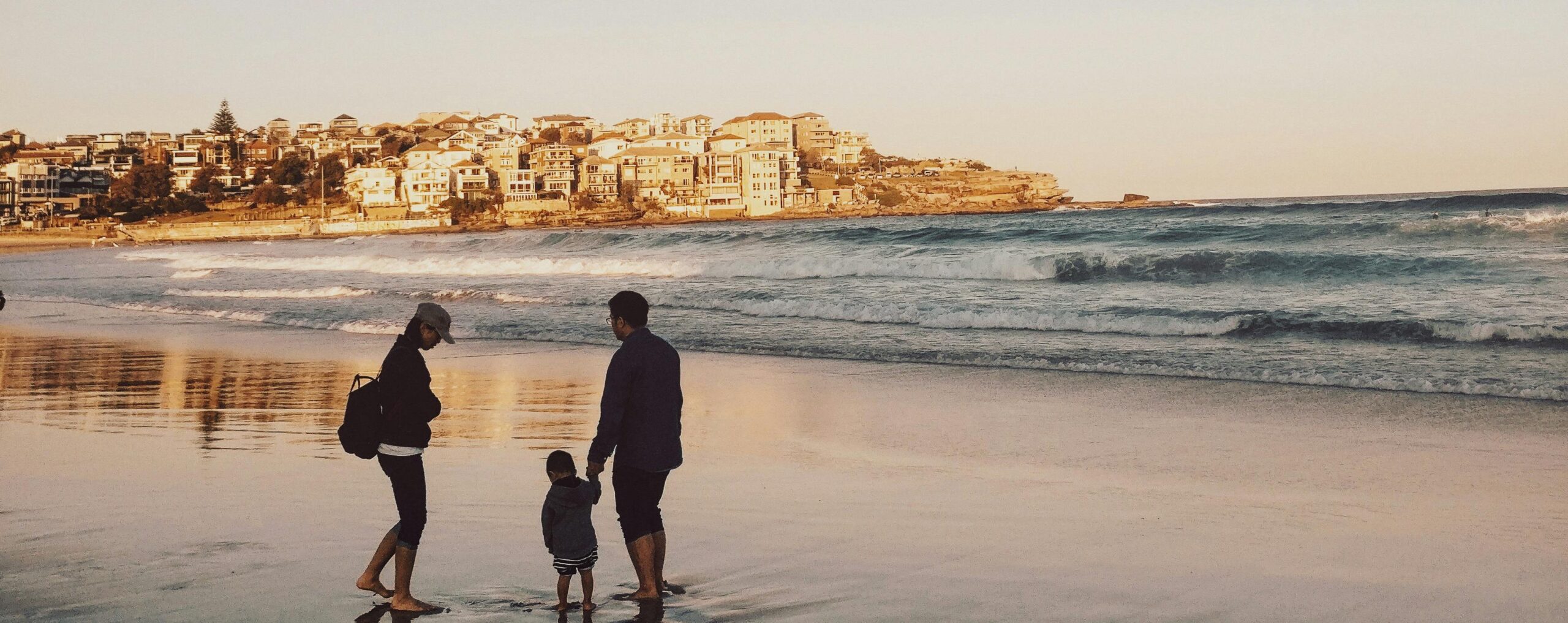 A woman, man and child are playing on a beach. In the background there is a view of a coastal town and a sunsetting sky.