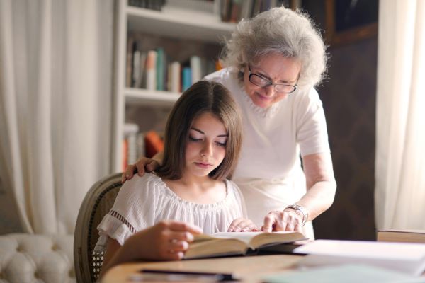 An older lady wearing a white top is helping a young teenage daughter read a book. In the background you can see a full bookshelf.