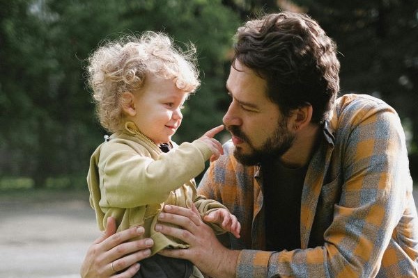 A man in an orange and grey check jumper is holding a young child who is touching him on the nose with its index finger. 