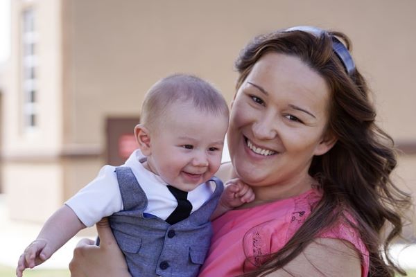 A woman in a pink dress is holding a small baby whilst both are smiling and looking into a camera.