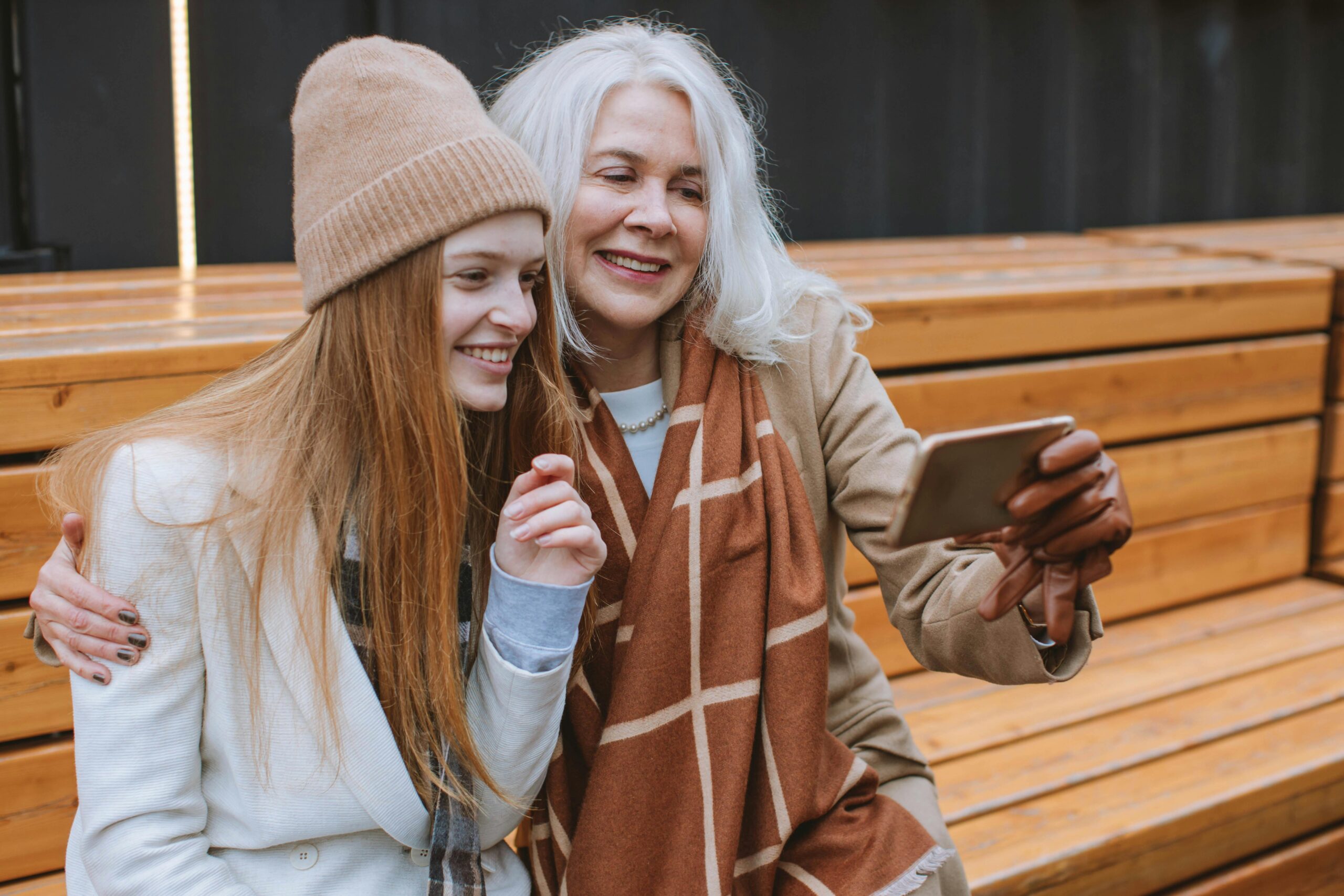 A teenage girl wearing a grey blazer and a light brown hat, sits on a wooden bench with an older woman who has a beige coat and dark brown patterned scarf. They are both smiling at a phone.