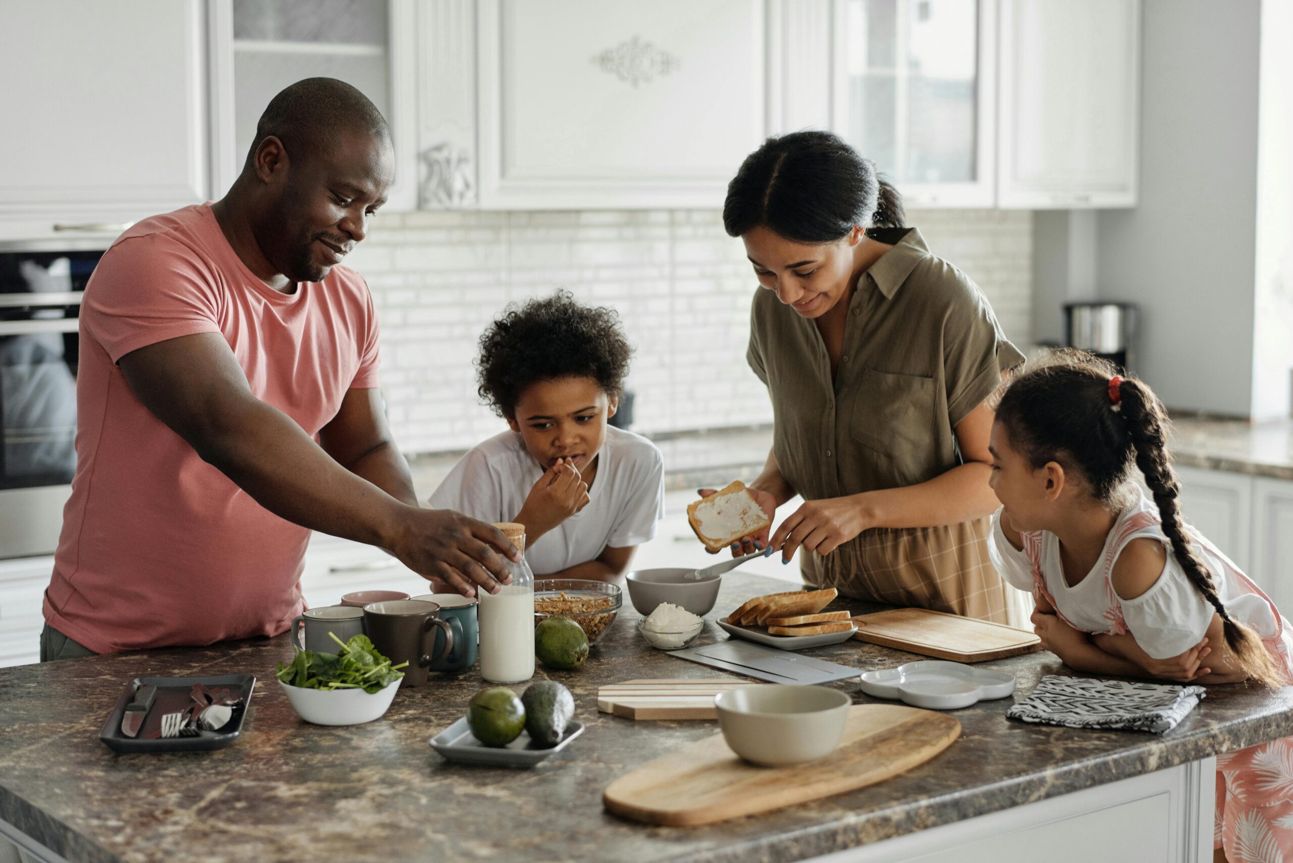 A family are stood around a kitchen island eating together. On the left there is a man in a pink t shirt lifting up a glass of milk. On his right is a boy watching on with a white top on learning on the island. Next to him is a woman in a dark top buttering bread and leaning over, on the far right of the image is a young girl peering to watch the woman.