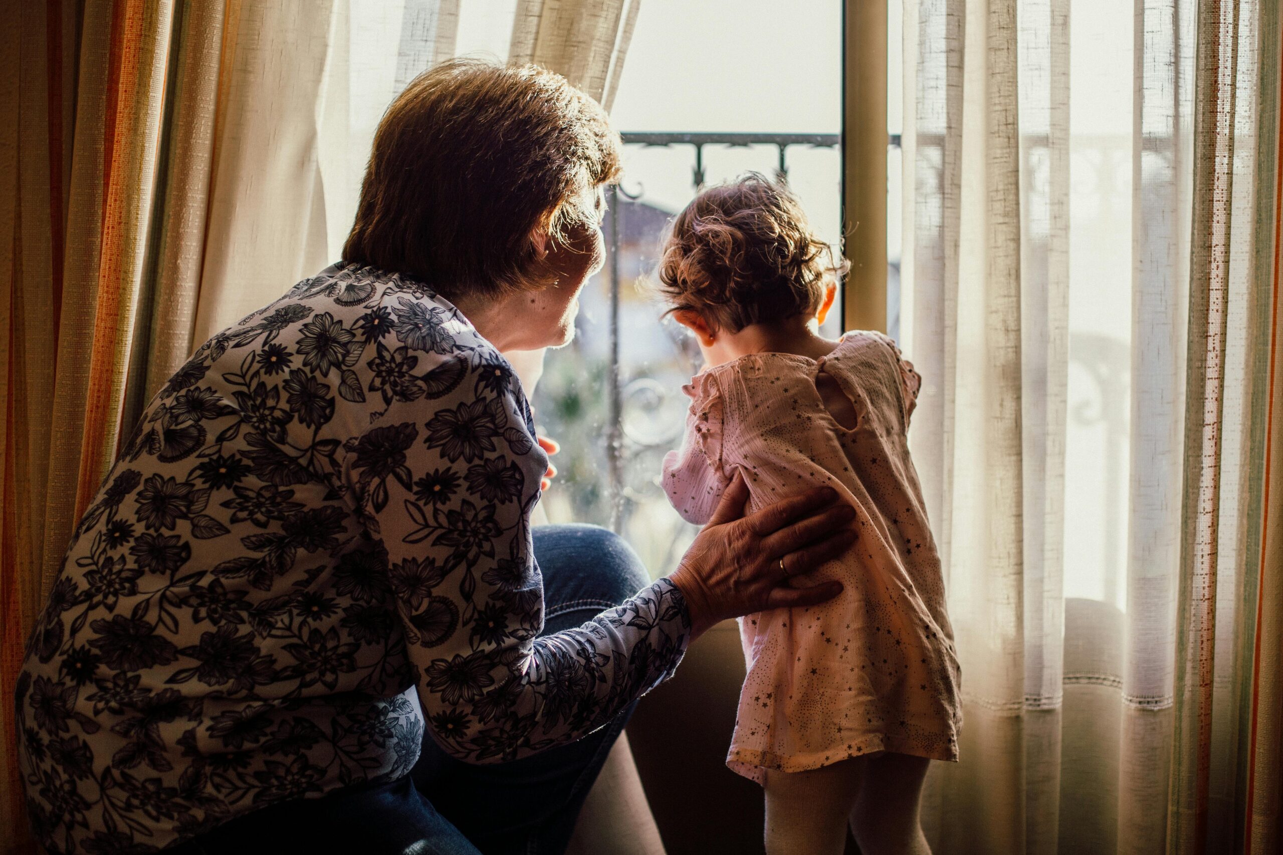 In the image there is an older woman and young child looking out of part drawn beige curtains. The older woman is on the left and is wearing a cream and blue floral patterned top. She has her right arm on the back of the young girl who is wearing a pink dress and leaning through the curtains.