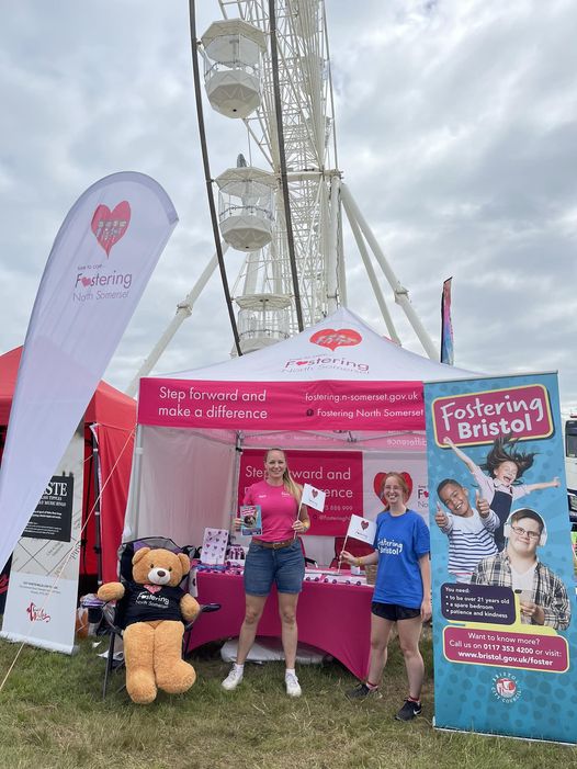 This photo shows a North Somerset Council and Bristol City Council Fostering Event. In front of a red and white marquee, there is a large teddy bear on a seat wearing a dark top. Next to them a woman stands in a bright red t shirt and denim shorts, and next to her is another woman in a blue top and denim shorts. On the right is a blue pull up banner advertising fostering for Bristol City Council. In the background there is a white ferris wheel.