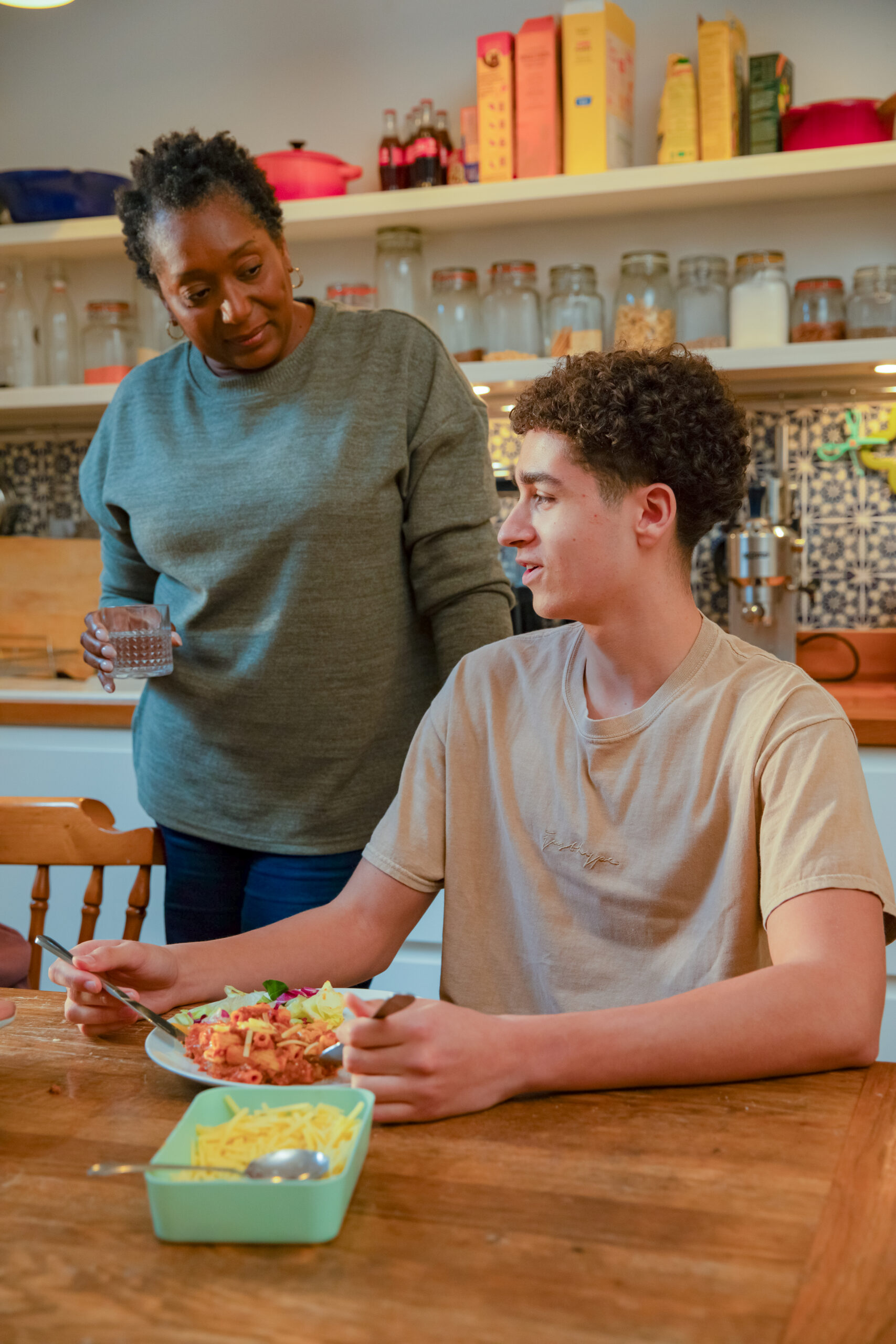 In a family kitchen setting a woman in a grey top stands next to a teenage boy who is sat at a wooden dining table in a beige top eating pasta and cheese.