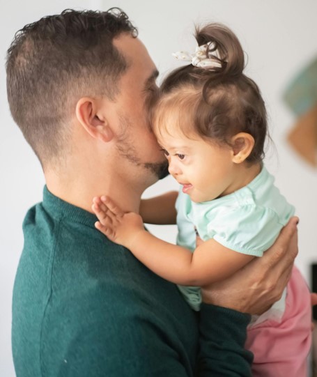 A man in a dark green top lifts up a young girl who is wearing pink shorts and has a light blue top on.