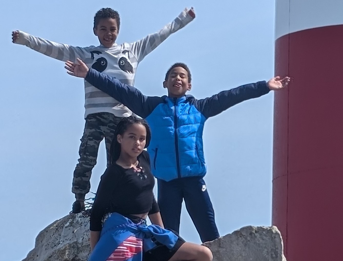 The image shows three children stood on a rock. Two young boys have their arms outstretched smiling to the sky, and a teenage girl looks at the camera.