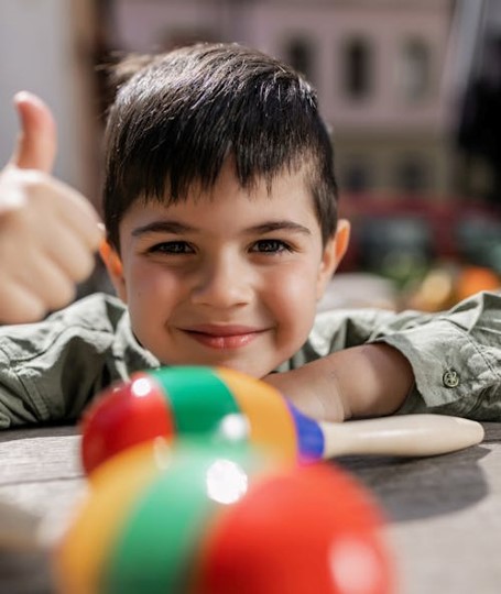 A child with dark hair holds their right thumb up whilst leaning on a bench. Children's' toys are in the foreground.