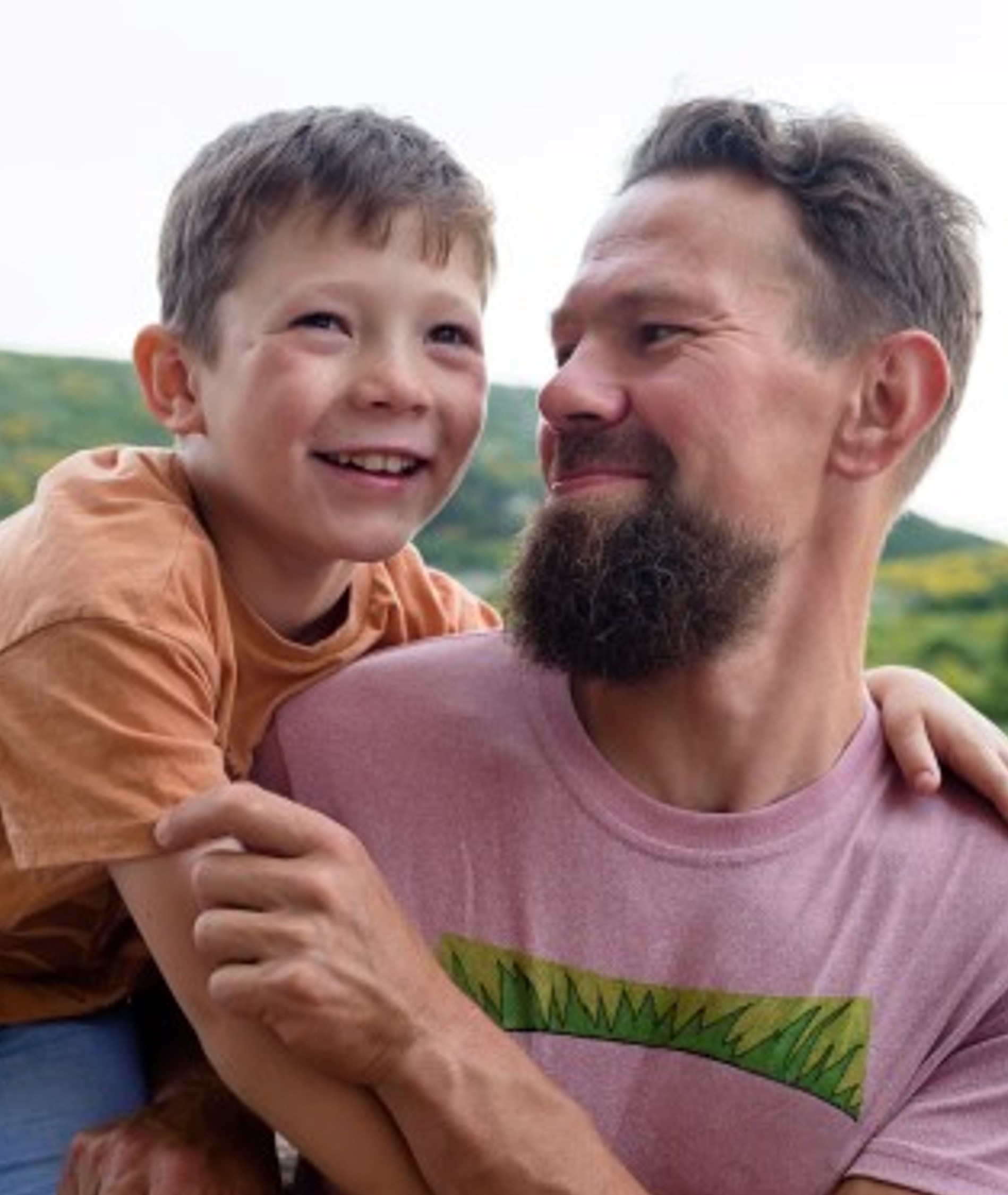 A man with dark hair and beard wearing a pink top smiles at a young boy who is on his shoulder.