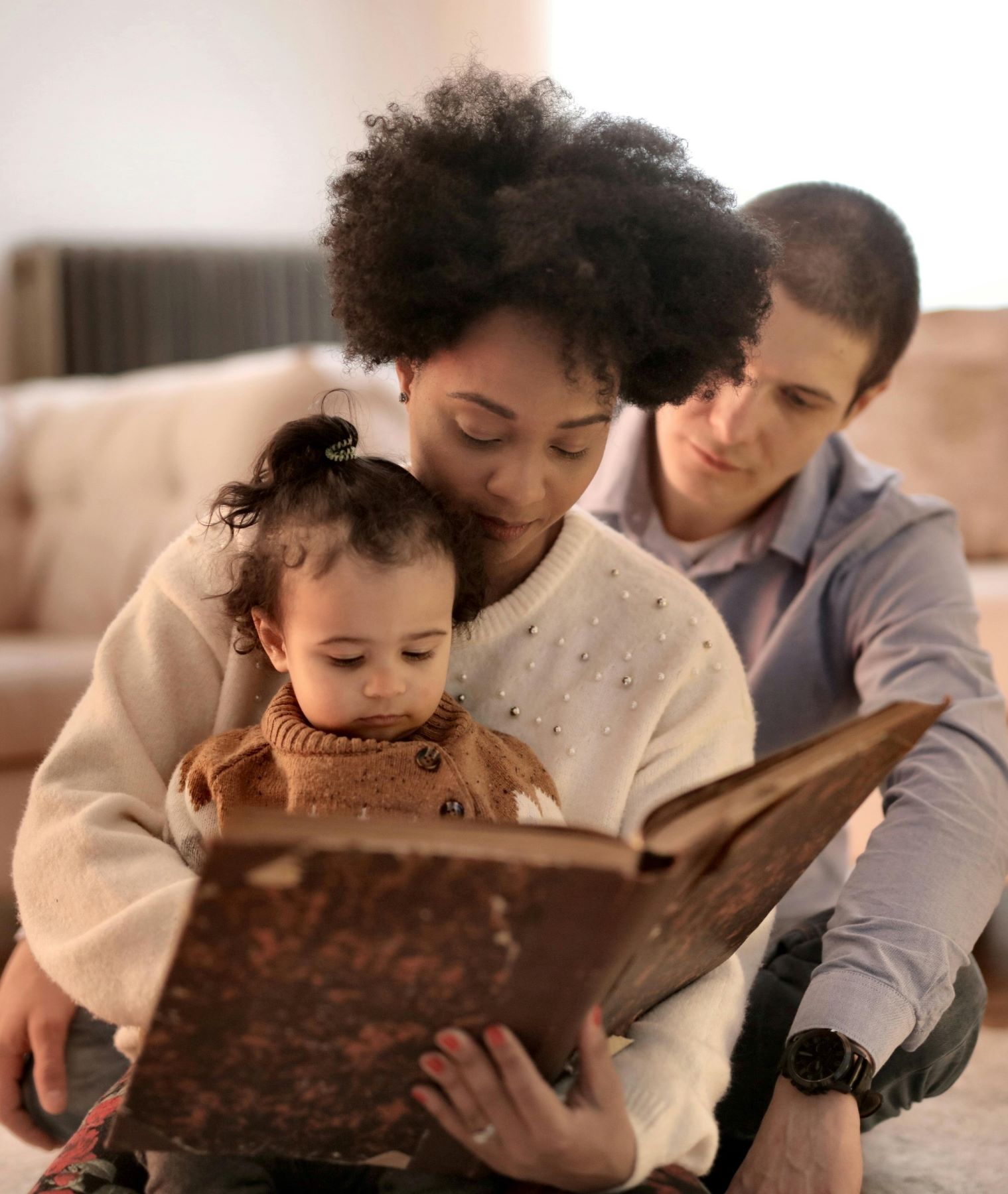 Sat on the floor with a sofa in the background you can see a man with short dark hair and a grey shirt on, infront of him is a woman with curly black hair and a white jumper. In his arms is a young girl with their hair in a bun and a brown jumper. The woman is reading to the child from a big book she is holding.