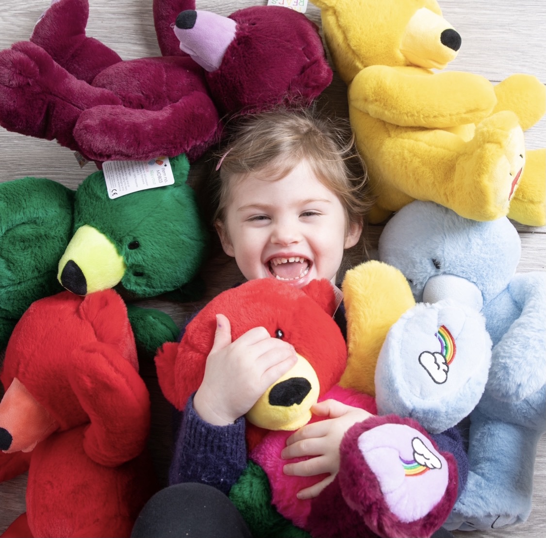 A young child is laughing and smiling at camera surrounded by a range of Mood Bear cuddly toys.