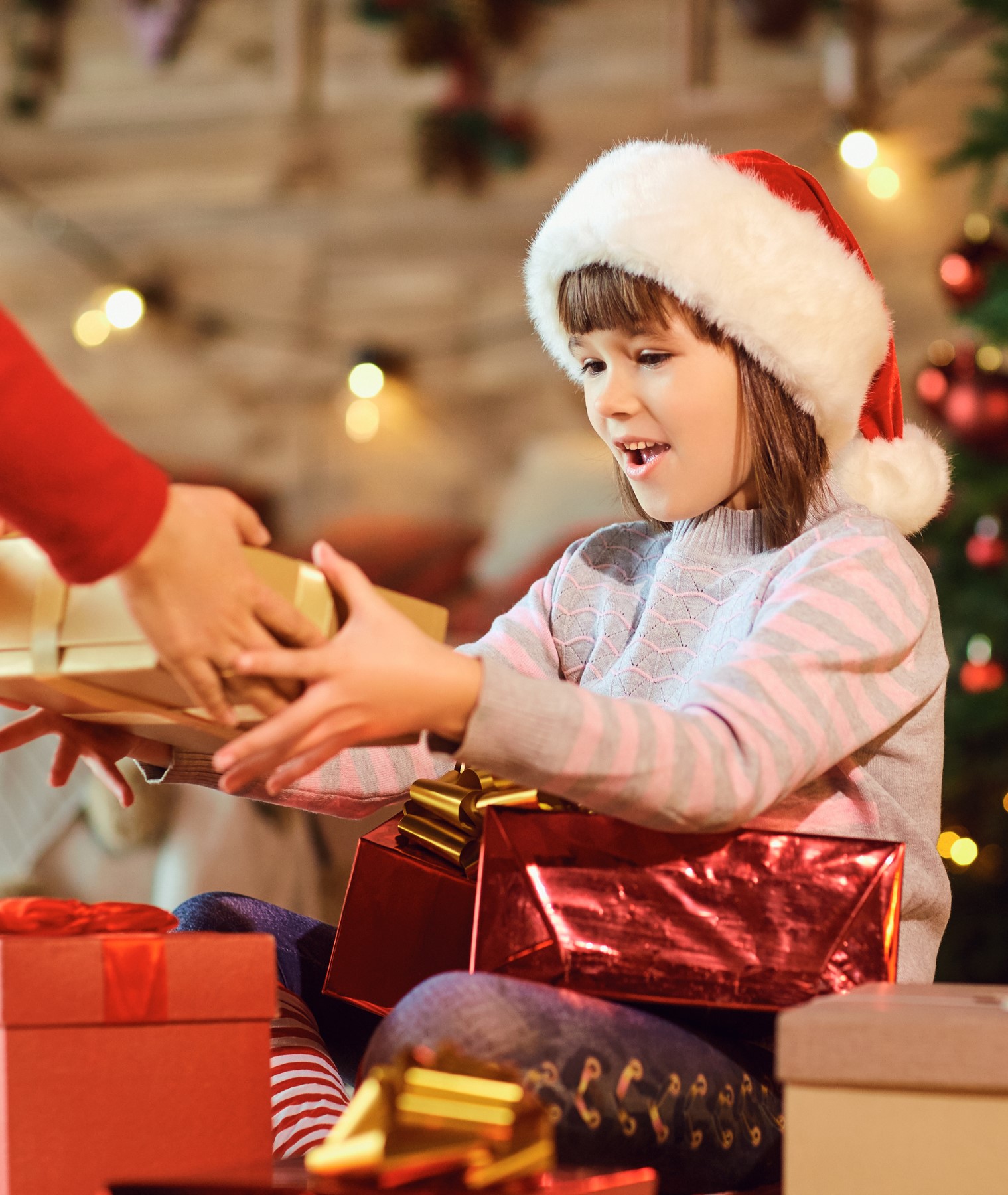 A young girl in a santa hat and grey and red pyjamas sits in front of a christmas tree and is handed a present.