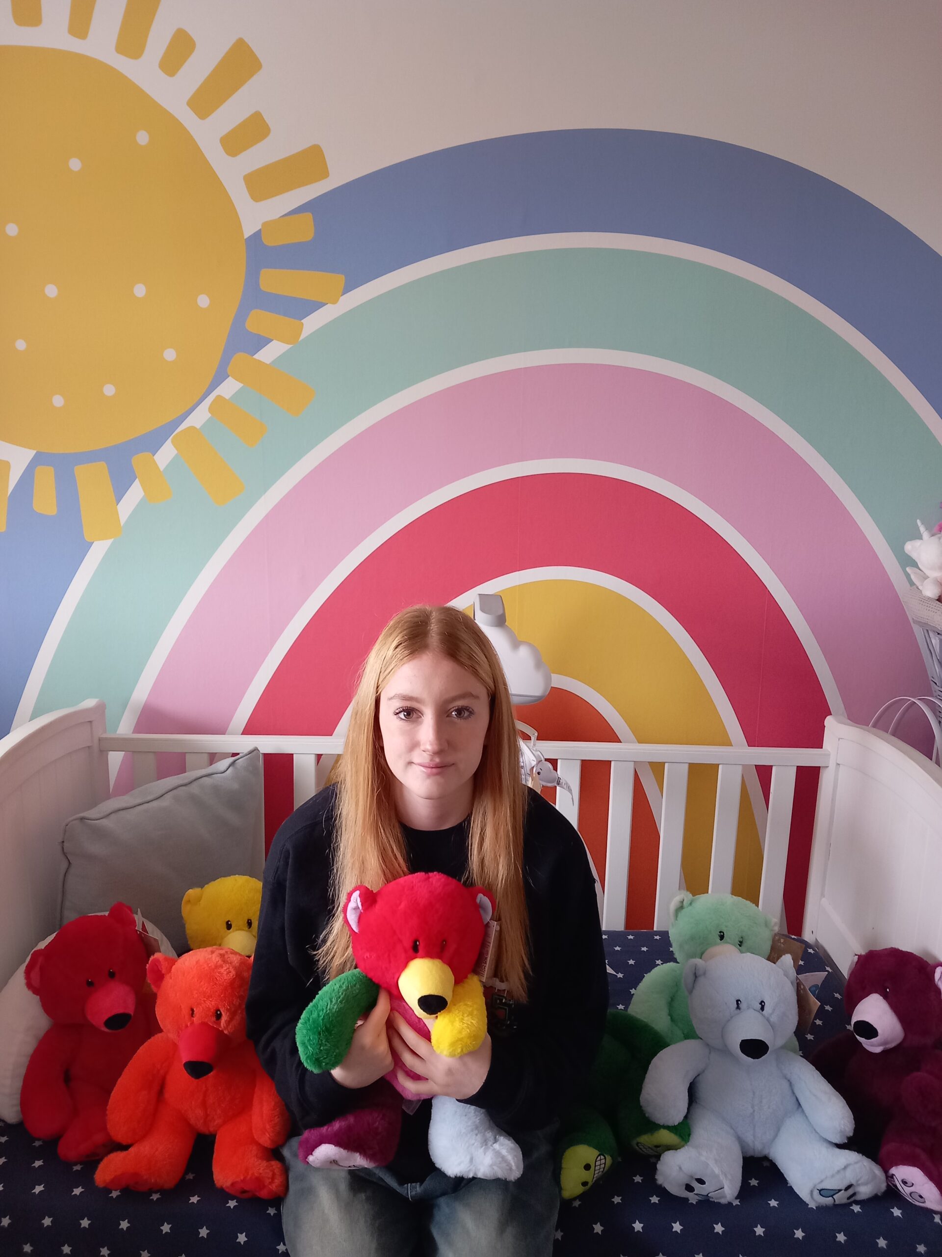 The umage shows a young teenage girl sat on a bed surrounded by mood bears. The background shows a rainbow painted wall.