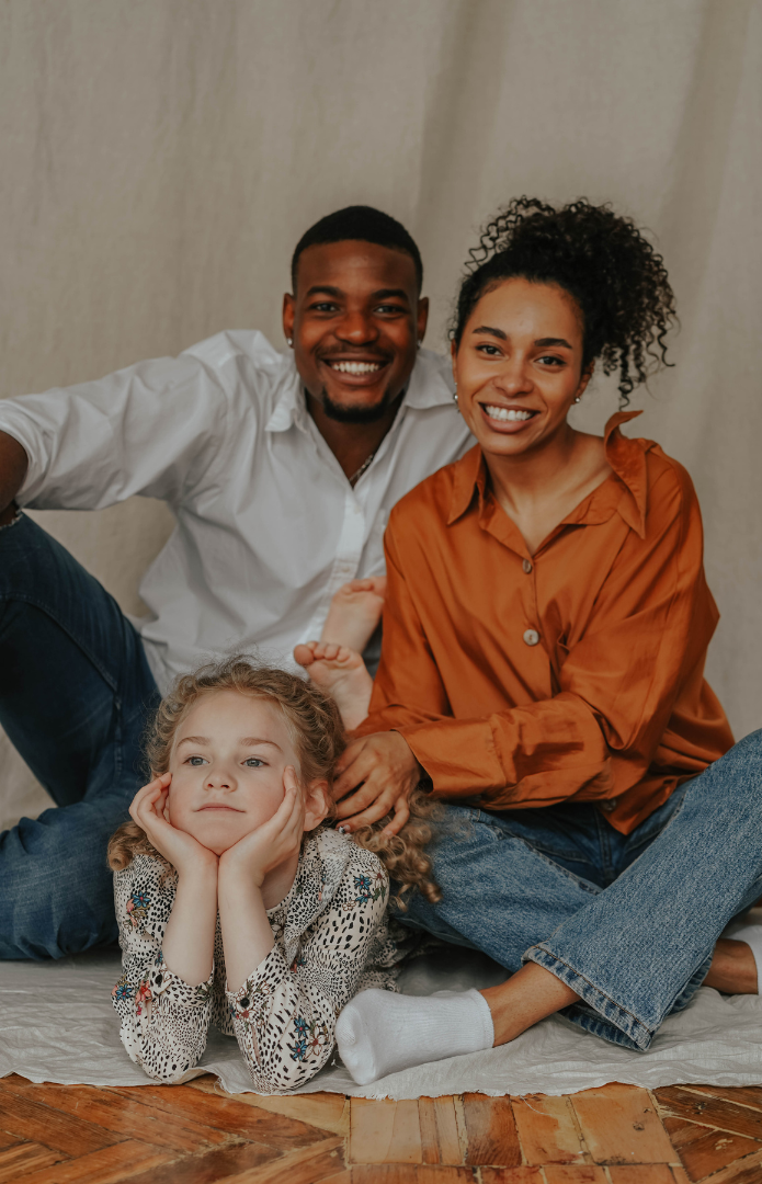 The image shows a man and woman smiling at camera sat on the floor, with a young girl lying in front of them with her hands on her chin