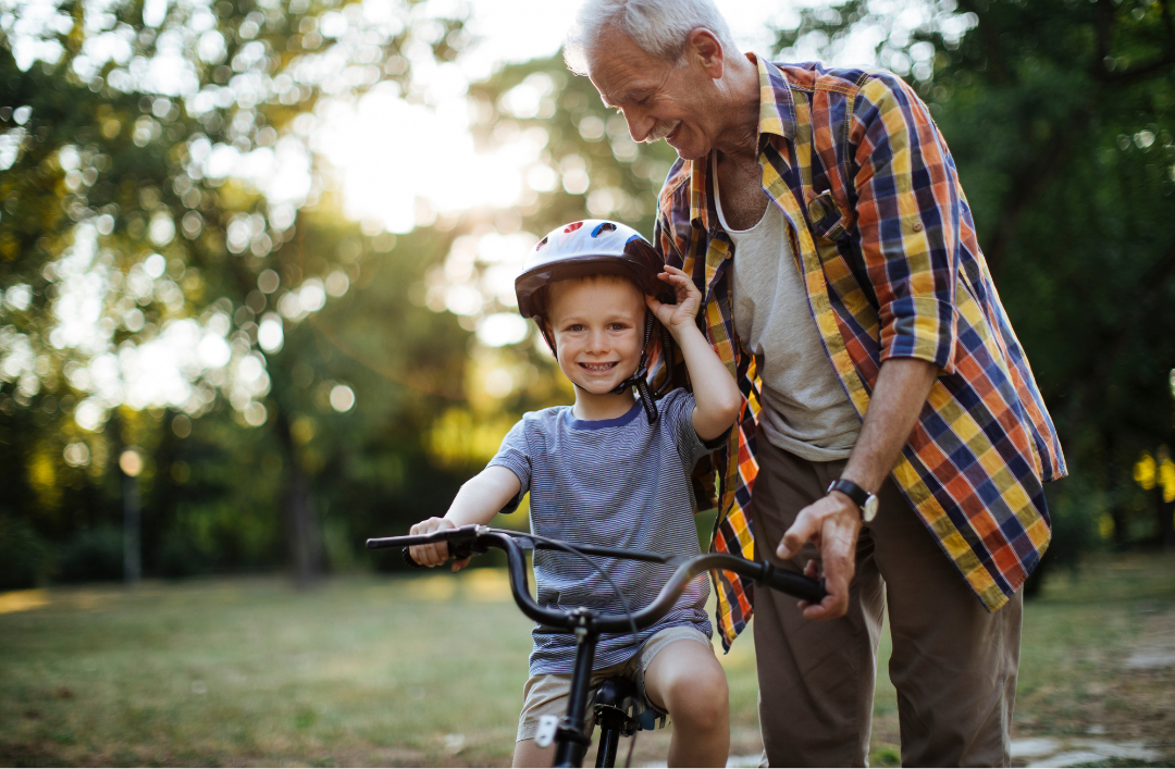 The image shows a young boy on a bike wearing a helmet. He's being held up by an older grey haired man.