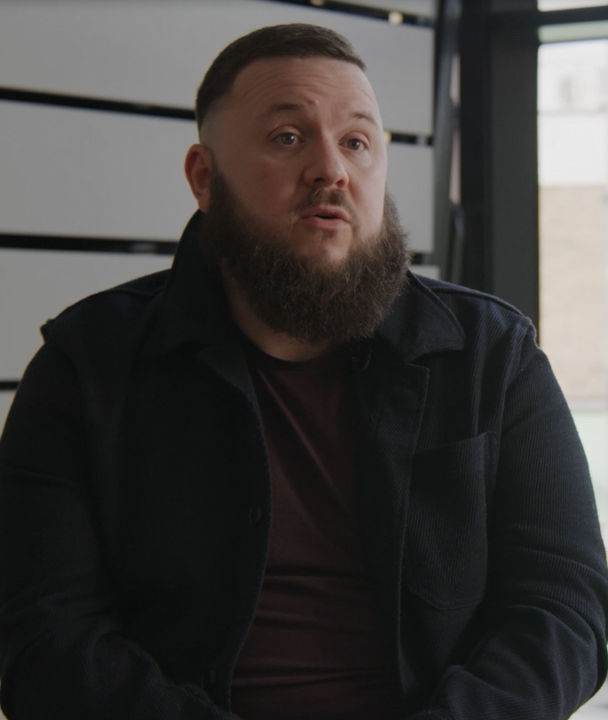 The image shows a man in a dark jacket with short brown hair and a large beard talking to camera