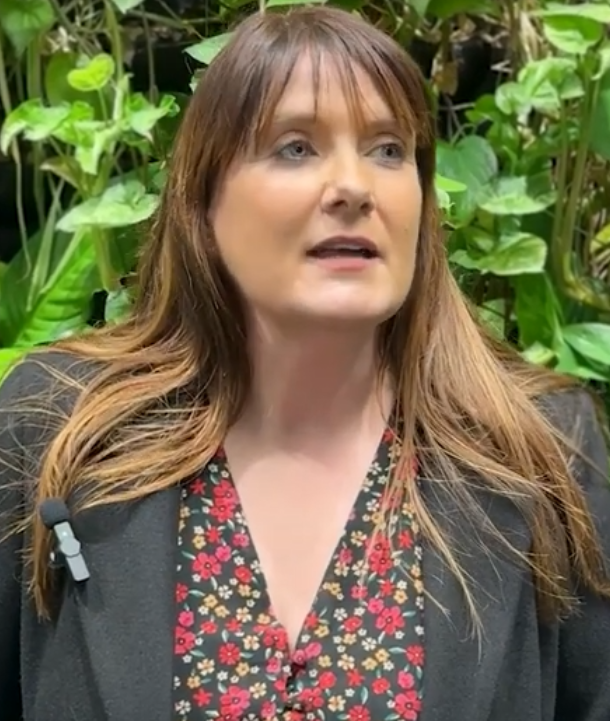 A woman stands in front of a leafy wall looking purposefully off camera